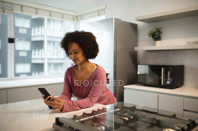 Vista frontal de una mujer de raza mixta relajándose en casa, de pie en la cocina apoyada en la encimera, usando un teléfono inteligente y sonriendo - foto de stock