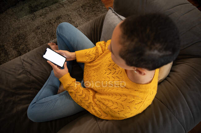 Vue grand angle d'une femme métissée se relaxant à la maison, assise sur un canapé avec les jambes croisées, à l'aide d'un smartphone — Photo de stock
