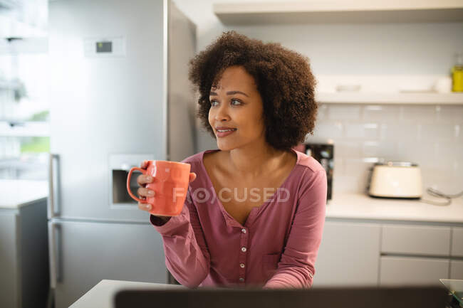 Vista frontal de una mujer de raza mixta relajándose en casa, de pie en la cocina apoyada en la encimera, mirando por la ventana y sosteniendo una taza de café y sonriendo - foto de stock