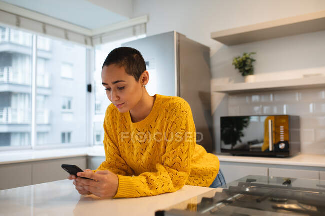 Вид спереду змішаної раси жінка з коротким волоссям, що розслабляється вдома, стоїть на кухні, спираючись на стільницю, використовуючи смартфон і посміхаючись — стокове фото