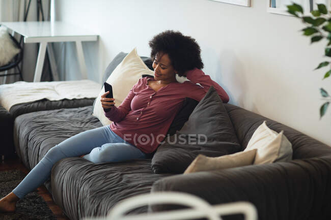 Seitenansicht einer Mischlingsfrau mit kurzen lockigen Haaren, die es sich zu Hause gemütlich macht, mit einem Smartphone auf einem Sofa sitzt und lächelt — Stockfoto