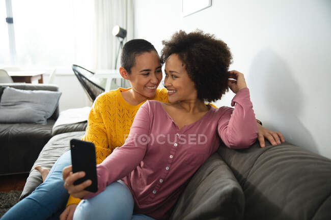 Вид спереду змішаної раси жіноча пара розслабляється вдома, сидячи на дивані, посміхаючись один одному, одна жінка тримає смартфон — стокове фото