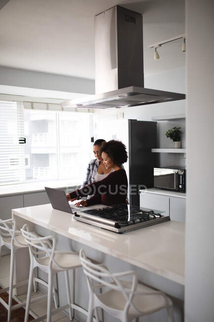 Vista lateral de cerca de una pareja femenina de raza mixta que se relaja en casa, de pie en la cocina usando un ordenador portátil juntos y sonriendo - foto de stock