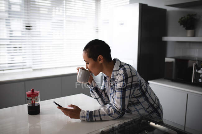 Vista lateral de una mujer de raza mixta con el pelo corto relajándose en casa, de pie en la cocina apoyada en la encimera, usando un teléfono inteligente y bebiendo una taza de café - foto de stock