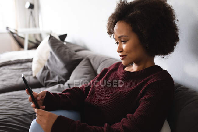 Вид сбоку на женщину смешанной расы, расслабляющуюся дома, сидящую на диване с помощью смартфона и улыбающуюся — стоковое фото