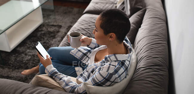 Високий кут зору змішаної раси жінка розслабляється вдома, сидячи на дивані за допомогою смартфона, тримаючи чашку кави і посміхаючись — стокове фото