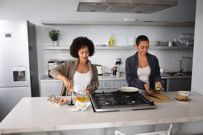Vista frontal de la pareja femenina de raza mixta relajándose en casa, de pie en la cocina preparando el desayuno juntos y sonriendo juntos - foto de stock