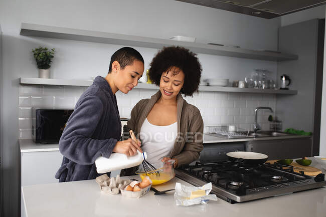 Вид спереду змішаної раси жіноча пара розслабляється вдома, стоячи на кухні, готуючи сніданок разом і посміхаючись, одна заливає молоко в миску, а інша змішує його з яйцями — стокове фото