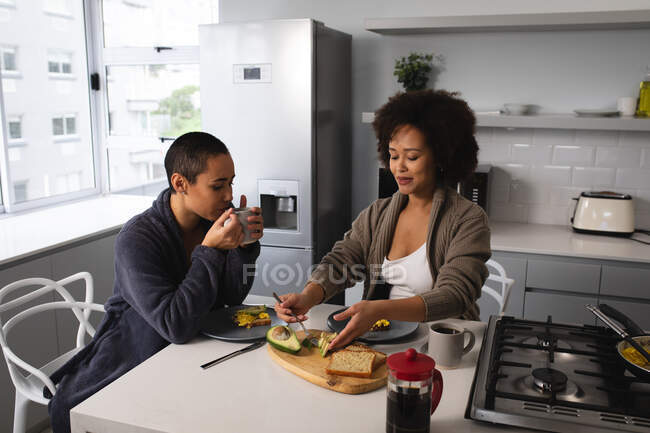 Вид спереду змішаної раси жіноча пара розслабляється вдома, сидячи на стільцях на кухонному острові, їсть сніданок і п'є каву — стокове фото