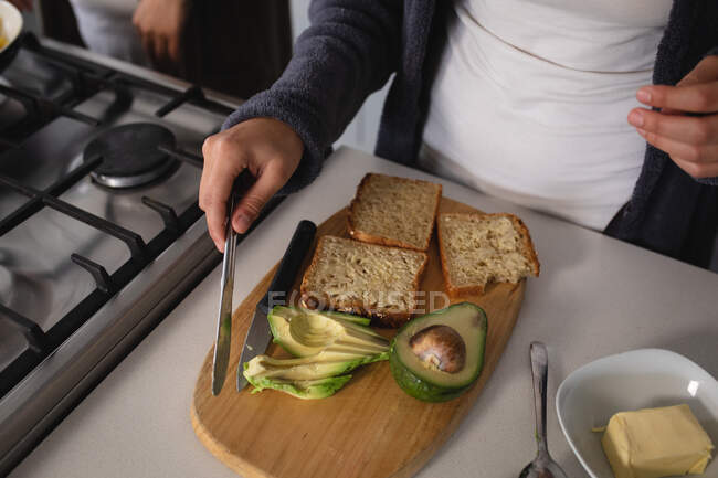 High angle front view mid section of woman relaxing at home, preparing breakfast in the kitchen, cutting avocado and oil bread on a chopping board — стоковое фото
