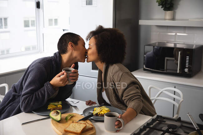 Vista frontal de una pareja femenina de raza mixta relajándose en casa sentada en sillas en la isla de la cocina desayunando y besando, sosteniendo tazas de café - foto de stock