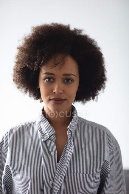 Retrato de perto de uma mulher de raça mista com cabelo encaracolado curto vestindo uma camisa de colarinho listrada aberta, preta e branca, olhando diretamente para a câmera em casa — Fotografia de Stock