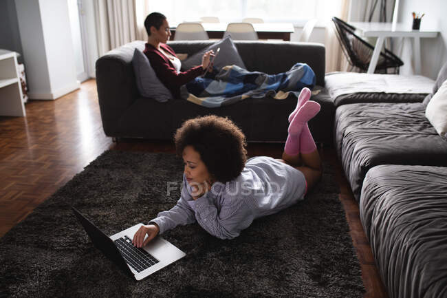 Вид збоку змішаної раси жіноча пара розслабляється вдома у вітальні, одна на дивані з ковдрою на ногах за допомогою смартфона, інша лежить на підлозі за допомогою портативного комп'ютера — стокове фото