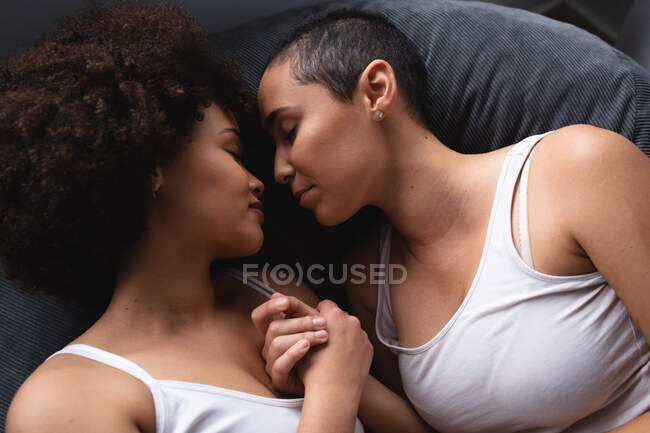 Высокий угол крупным планом вид смешанной расовой пары женщин, отдыхающих дома в спальне вместе по утрам, лежащих лицом друг к другу с закрытыми глазами и держащихся за руки — стоковое фото