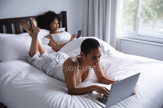 Seitenansicht eines gemischten Rennfahrerpaares, das es sich morgens zu Hause im Schlafzimmer gemütlich macht, wobei die eine mit einem Smartphone im Bett sitzt und die andere mit einem Laptop im Bett liegt. — Stockfoto