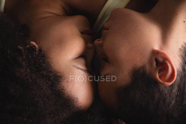 Overhead shot de una pareja femenina de raza mixta relajándose en casa, acostados en la cama dormidos con sus caras tocando - foto de stock