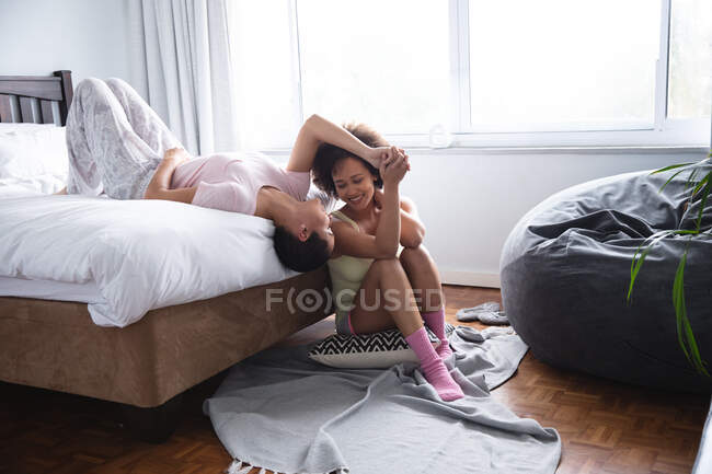 Vista frontal de una pareja femenina de raza mixta que se relaja en casa en el dormitorio por la mañana, una acostada boca arriba en la cama y la otra sentada junto a ella en el suelo, sonriéndose la una a la otra y tomándose de la mano - foto de stock