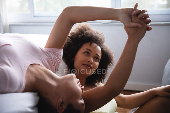 Vista frontal de cerca de una pareja femenina de raza mixta que se relaja en casa en el dormitorio por la mañana, una acostada boca arriba en la cama y la otra sentada junto a ella en el suelo, cogida de la mano y sonriendo - foto de stock
