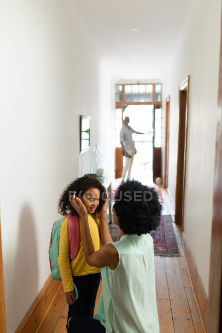 Vue de face d'une jeune afro-américaine debout dans le couloir à la maison portant un sac à dos et souriant, avec sa mère agenouillée à côté d'elle disant au revoir et son père debout près de la porte d'entrée ouverte en arrière-plan — Photo de stock