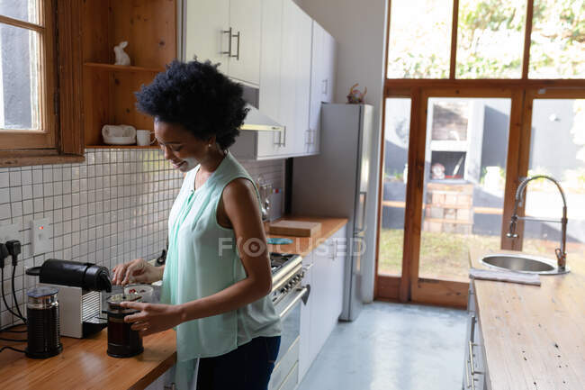 Придивіться до привабливої афро - американської жінки вдома, яка стоїть на кухні і готує каву в кафетерії. Вона посміхається і насолоджується вихідними.. — стокове фото