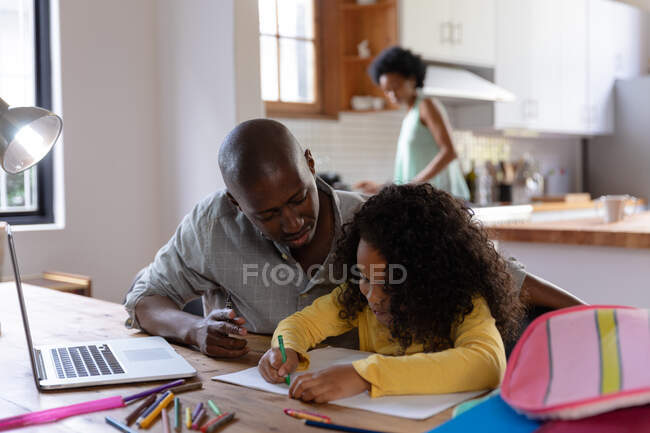 Передній погляд на афроамериканця вдома, який сидів за столом зі своєю молодшою донькою і дивився на малюнок у шкільній книжці, ноутбук на столі перед ним, з матір'ю, що стояла на кухні на задньому плані. — стокове фото