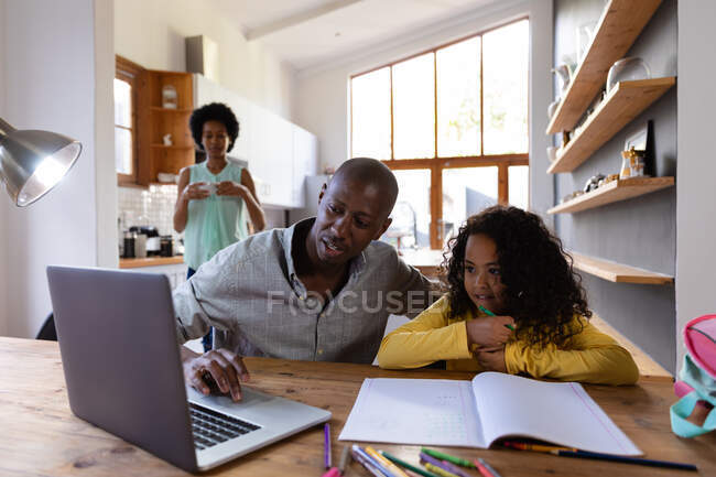 Vorderansicht eines afroamerikanischen Mannes zu Hause, der mit seiner kleinen Tochter an einem Tisch sitzt und gemeinsam auf einen Laptop schaut, ein Schulbuch auf dem Tisch vor sich, im Hintergrund steht die Mutter in der Küche — Stockfoto