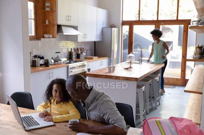 Vista de alto ângulo de um homem afro-americano em casa, sentado em uma mesa com sua filha olhando para um computador portátil juntos e conversando, com a mãe de pé na cozinha no fundo — Fotografia de Stock