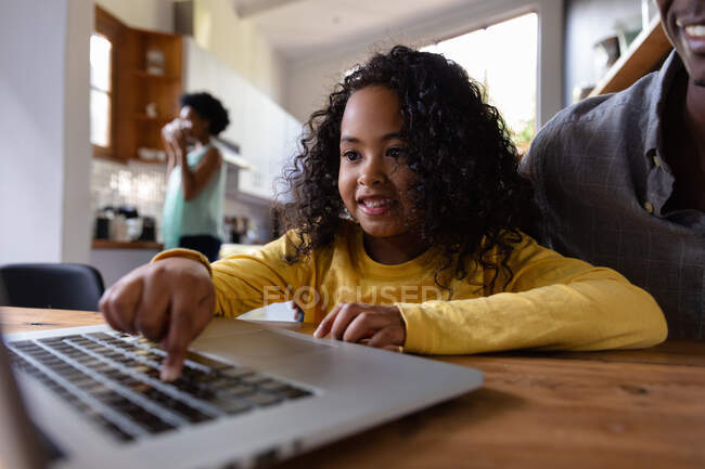 Front view close up of a young African American girl at home, sitting at a table with her father looking at a laptop computer together, the girl pressing the computer keyboard and smiling, with the mother standing in the kitchen in the background — Stock Photo