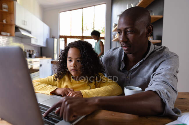 Front view close up of a young African American girl at home, sitting at a table with her father looking at a laptop computer together, the father pressing the computer keyboard and smiling, with the mother standing in the kitchen in the background — Stock Photo
