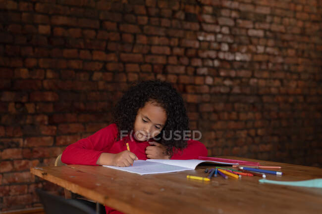 Передній погляд на молоду афроамериканку вдома, яка сидить за обіднім столом за допомогою олівців і виконує домашнє завдання, оголену цегляну стіну на задньому плані. — стокове фото