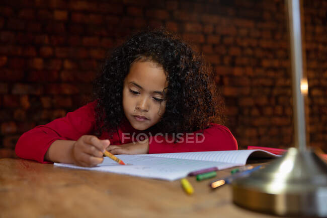 Vista frontale da vicino di una giovane ragazza afroamericana a casa, seduta a tavola a fare i compiti, colorata con un pastello in un libro scolastico — Foto stock