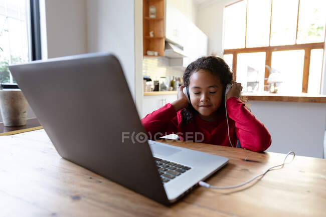 Вид спереди на молодую афроамериканку, сидящую дома за обеденным столом, слушающую с закрытыми глазами и включенными наушниками, подключенную к ноутбуку на столе перед ней и улыбающуюся — стоковое фото