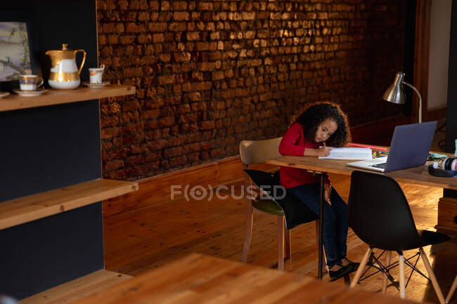 Vista lateral de una joven afroamericana en casa, sentada en la mesa de la cena haciendo sus deberes, escribiendo en un libro escolar con un portátil en la mesa frente a ella - foto de stock