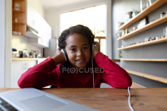 Vue de face gros plan d'une jeune afro-américaine à la maison, assise à la table du dîner, écoutant avec un casque, branchée sur un ordinateur portable sur la table devant elle, regardant vers la caméra et souriant — Photo de stock