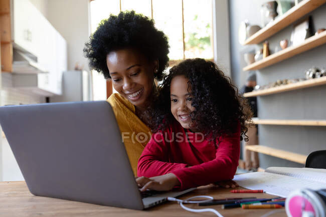 Передній погляд закриває будинок молодої афроамериканської дівчини, яка сидить за столом зі своєю матір'ю і дивиться на портативний комп'ютер разом, дочка натискає на клавіатуру комп'ютера і обидва посміхаються — стокове фото