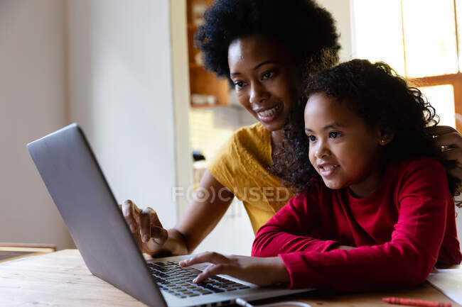 Vista lateral de cerca de una joven afroamericana en casa, sentada en una mesa con su madre mirando un ordenador portátil juntos, la hija presionando el teclado del ordenador y ambos sonriendo - foto de stock