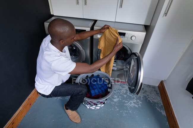 Vista de alto ángulo de un hombre afroamericano en casa, arrodillado y sacando la ropa de una lavadora - foto de stock