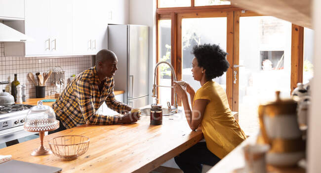 Vista lateral de una pareja afroamericana en casa en la cocina, sentada a cada lado de la isla de la cocina, tomando café y hablando - foto de stock
