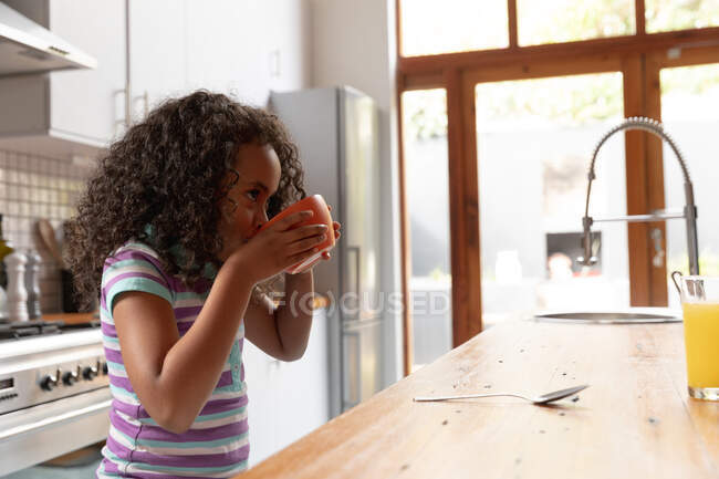 Side view of a young African American girl at home in the kitchen, standing at the kitchen island eating breakfast cereal from the bowl — Stock Photo