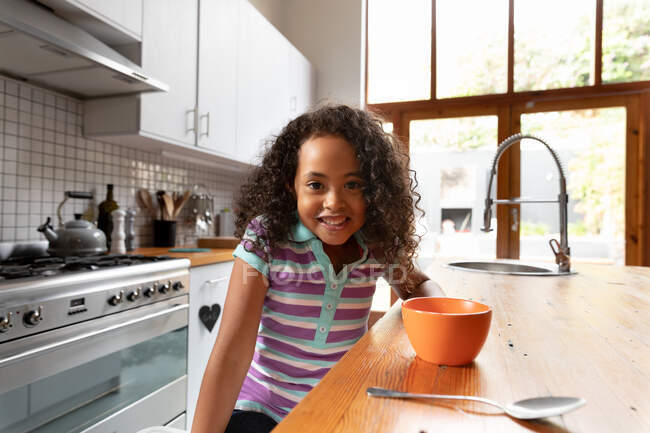 Portrait of a young African American girl at home in the kitchen, sitting at the kitchen island in the morning, with a bowl of breakfast cereal and a spoon in front of her, smiling to camera — Stock Photo