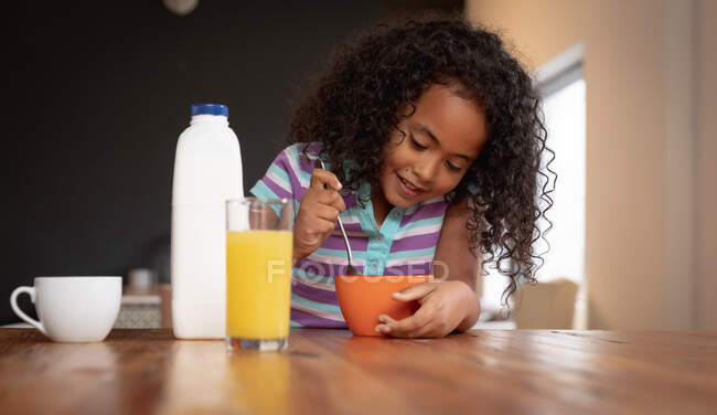 Vue de face gros plan d'une jeune afro-américaine à la maison dans la cuisine, assise à une table mangeant des céréales pour le petit déjeuner et souriant — Photo de stock