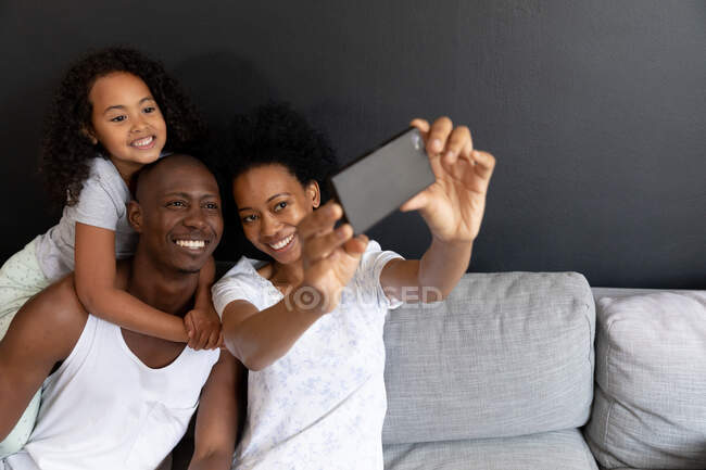 Frontansicht eines afroamerikanischen Paares und ihrer kleinen Tochter, die es sich morgens im Wohnzimmer gemütlich machen. Das Paar sitzt auf dem Sofa, die Tochter steht hinter ihrem Vater, die Hände Arme um seinen Hals. — Stockfoto