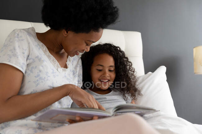 Vue de face gros plan d'une femme afro-américaine et de sa jeune fille se relaxant dans la chambre, s'asseyant au lit et lisant un livre ensemble — Photo de stock