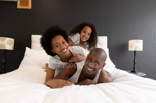 Front view of an African American couple and their young daughter relaxing in the bedroom, lying in a pile on the bed together and looking to camera smiling — Stock Photo