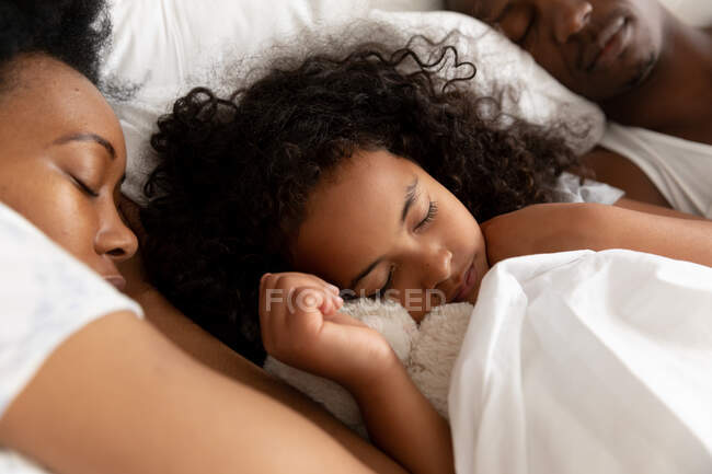 Vue de face rapprochée d'une jeune afro-américaine couchée au lit entre ses parents endormis. Ils se détendent ensemble. — Photo de stock