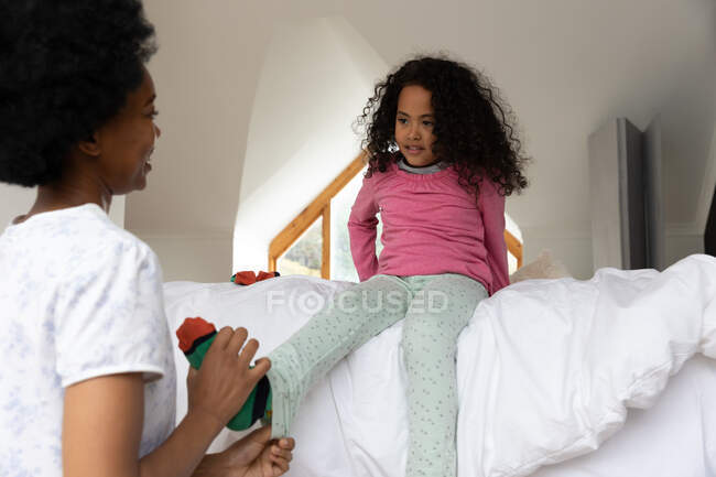 Vista frontale di una giovane ragazza afroamericana in camera da letto, seduta sul letto, con la madre in primo piano, che la aiuta a vestirsi — Foto stock