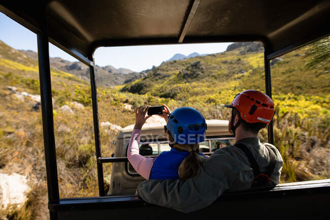 Rear view of Caucasian couple enjoying time in nature together, in zip lining equipment sitting in a car, the woman taking photos with smartphone on a sunny day in mountains — Stock Photo