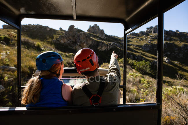 Rear view of Caucasian couple enjoying time in nature together, in zip lining equipment sitting in a car, the man is pointing with his finger, on a sunny day in mountains — Stock Photo