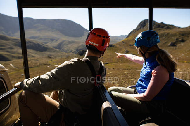 Rückansicht eines kaukasischen Paares, das die Zeit in der Natur zusammen genießt, in Reißverschlussausrüstung im Auto sitzend, zeigt die Frau mit dem Finger auf einen sonnigen Tag in den Bergen — Stockfoto