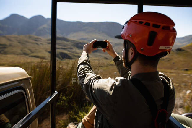 Side view of Caucasian man enjoying time in nature, in zip lining equipment sitting in a car, taking photos with smartphone on a sunny day in mountains — Stock Photo